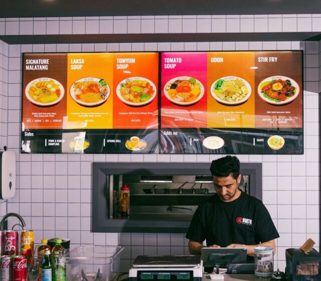 A North Hot Pot team member at the ordering counter, beneath the lit broth menu boards